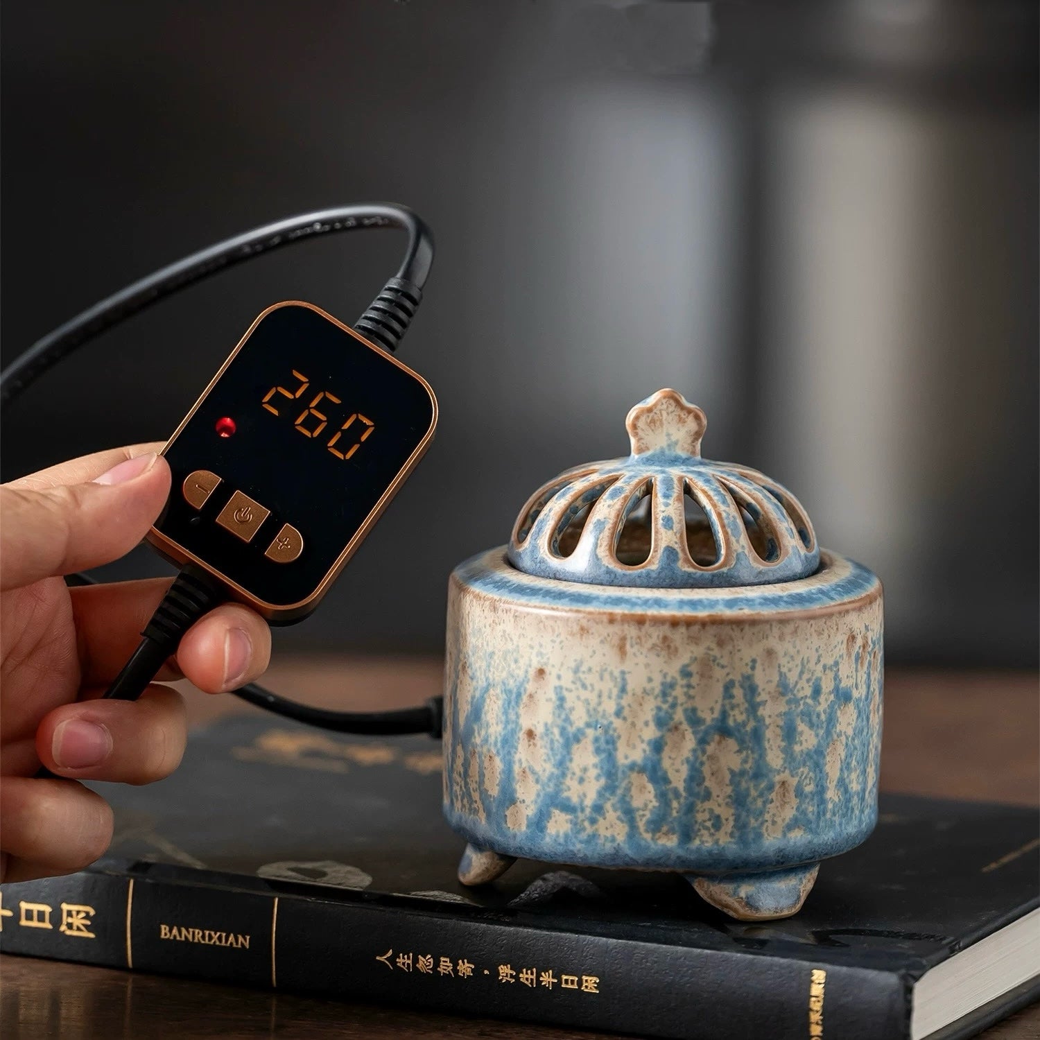 Hand holding a digital temperature controller set to 260 beside a handmade blue ceramic electric incense burner placed on stacked books.