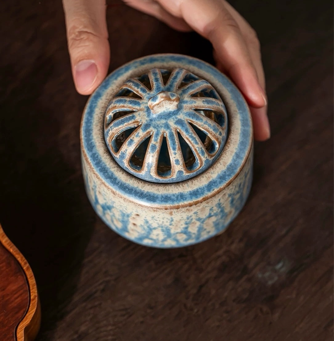 Handmade ceramic electric incense burner with the vented lid on, held by two hands on a wooden table for aromatherapy use.