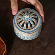 Handmade ceramic electric incense burner with the vented lid on, held by two hands on a wooden table for aromatherapy use.