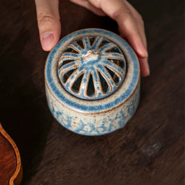 Handmade ceramic electric incense burner with the vented lid on, held by two hands on a wooden table for aromatherapy use.
