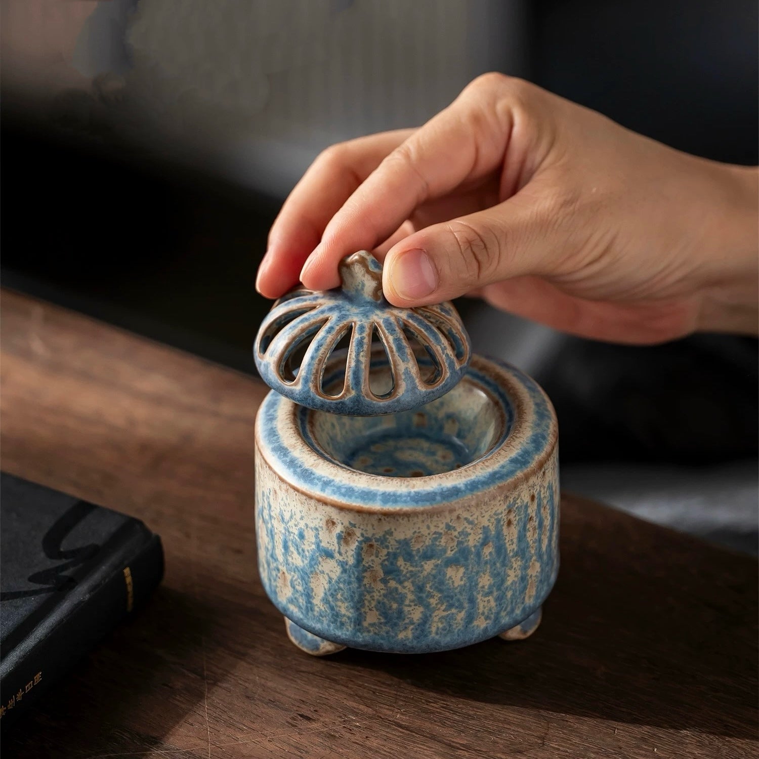 Hand lifting the vented lid off a handmade blue ceramic electric incense burner, showing the inner bowl on a wooden table.