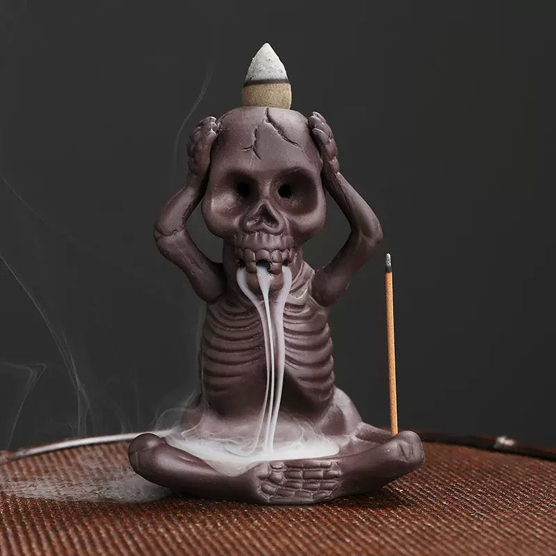 A seated skeleton incense burner covers its ears in a “hear no evil” pose, with a backflow cone on top releasing smoke through the mouth and a slim stick incense holder standing beside it.