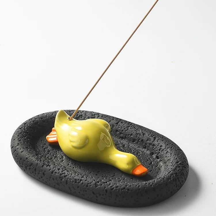High-angle studio shot of a yellow ceramic duck incense holder on a black, porous oval tray. The image clearly shows the rough, stone-like texture of the black base and the incense stick inserted in the duck.