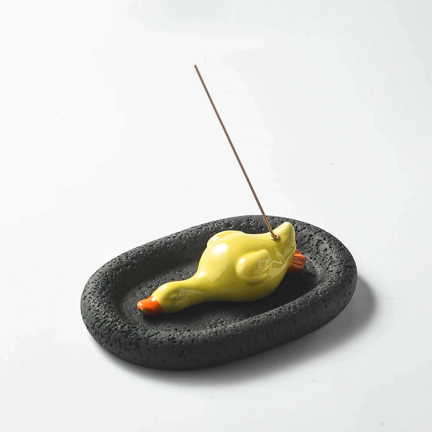 A studio shot of the assembled incense set featuring a yellow ceramic duck on a black, porous oval tray. An incense stick is upright in the holder, showcasing the striking color contrast between the bright duck and the dark base.