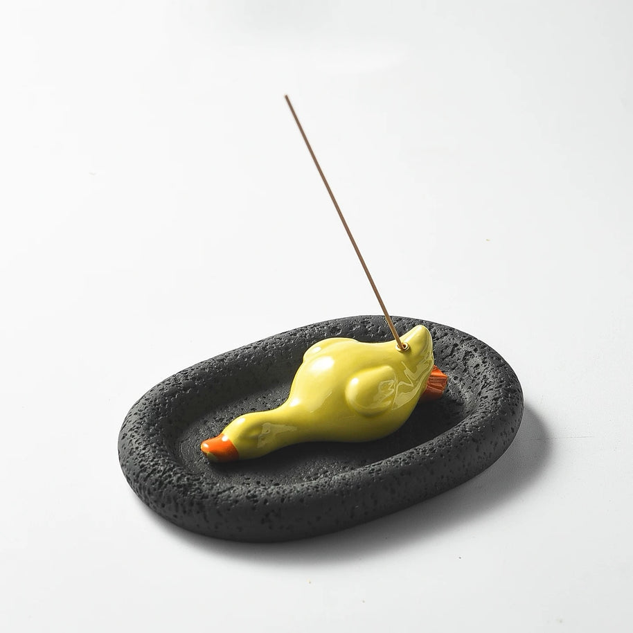 A studio shot of the assembled incense set featuring a yellow ceramic duck on a black, porous oval tray. An incense stick is upright in the holder, showcasing the striking color contrast between the bright duck and the dark base.