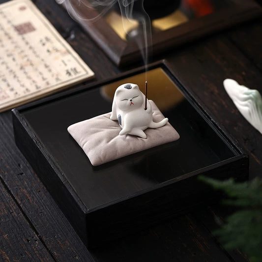 White ceramic cat incense holder on a cushion with a lit stick and rising smoke; Japanese zen desk decor.