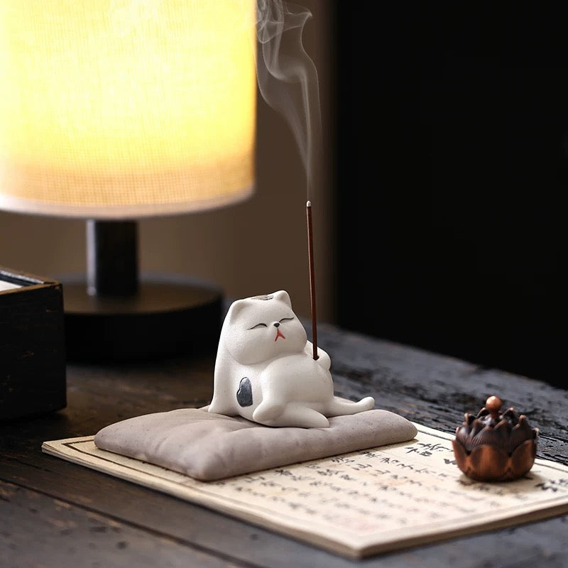 White ceramic sleeping cat incense holder on a cushion, lit stick with rising smoke, on calligraphy paper with a warm lamp in the background.