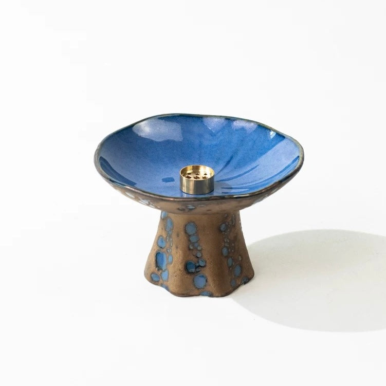 Handmade Japanese wabi-sabi style pedestal ceramic incense holder with a glossy blue dish and brown base with blue spots, featuring a brass incense insert on a white background.