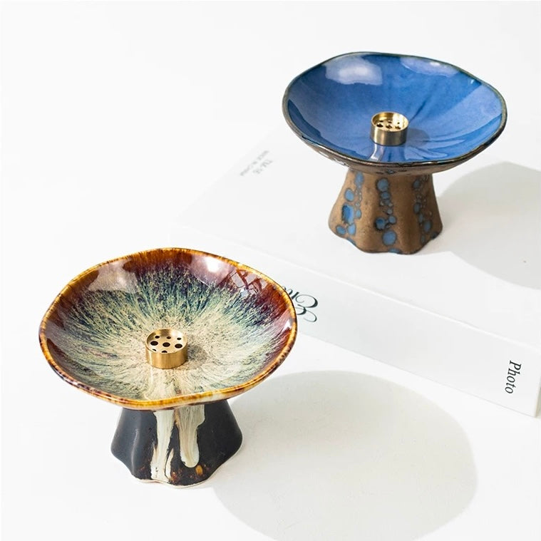 Two Japanese wabi-sabi style pedestal ceramic incense holders on a white background, one with a brown reactive glaze dish and one with a glossy blue dish on a brown base, both featuring brass incense inserts.