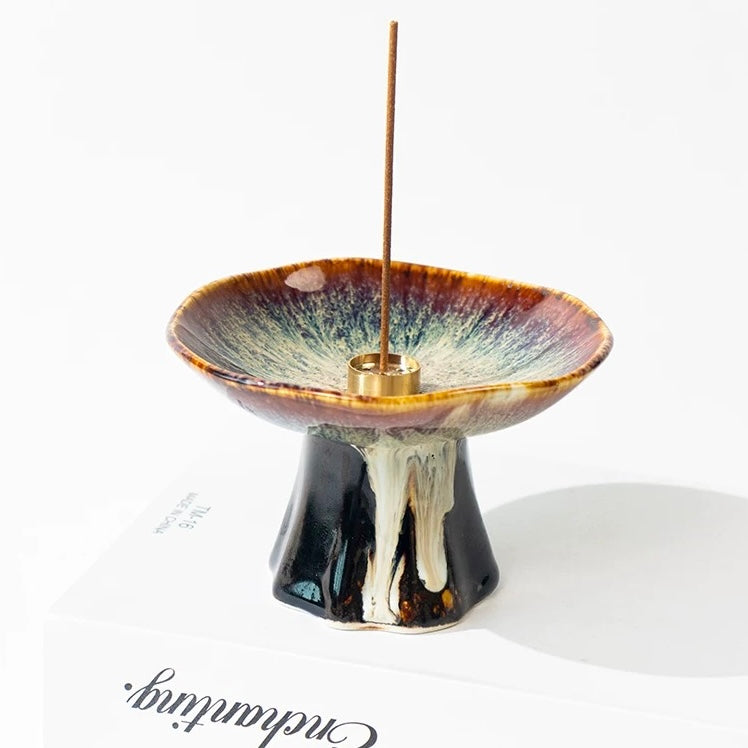 Japanese wabi-sabi style brown pedestal ceramic incense holder with reactive glaze and a brass insert holding a vertical incense stick, displayed on a white background.