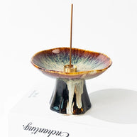 Japanese wabi-sabi style brown pedestal ceramic incense holder with reactive glaze and a brass insert holding a vertical incense stick, displayed on a white background.
