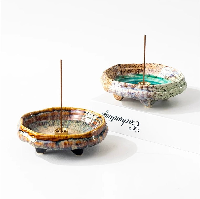 Two handmade Japanese wabi-sabi style ceramic incense holders, one green and one brown, each with a brass incense insert and incense stick, displayed on a white background.
