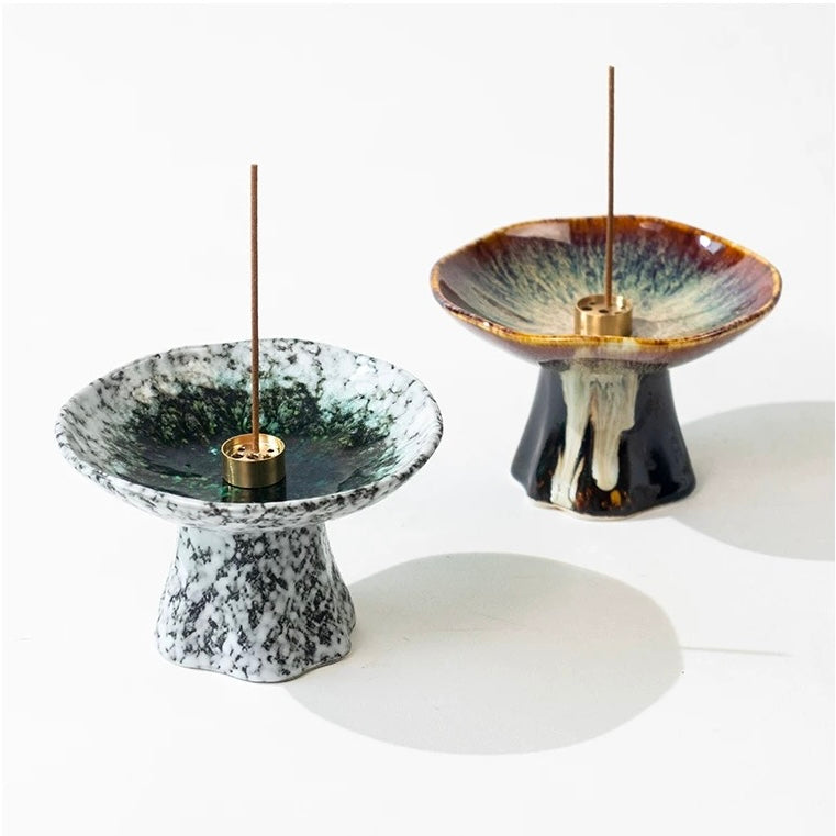 Two handmade Japanese wabi-sabi style pedestal ceramic incense holders with brass incense inserts and burning incense sticks, one white speckled and one brown with flowing glaze, on a white background.
