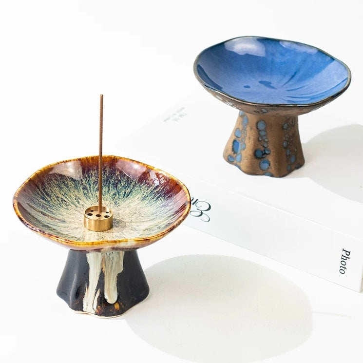 Two Japanese wabi-sabi style pedestal ceramic incense holders, one with a reactive brown-and-cream glaze and incense stick, and one with a glossy blue dish on a brown base, displayed on a white background.