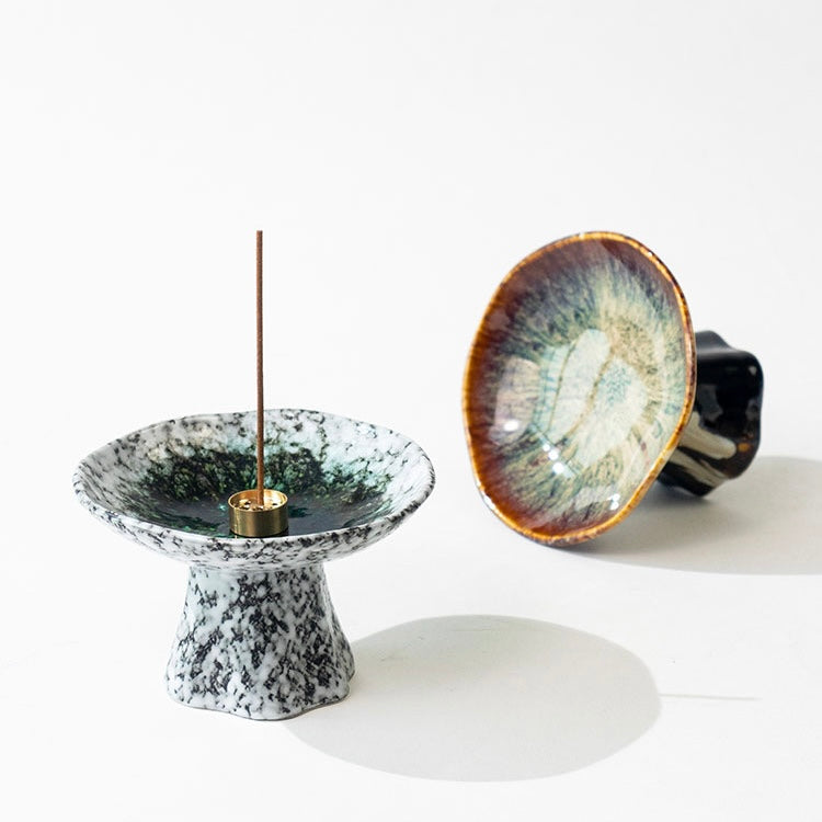 Japanese wabi-sabi style pedestal ceramic incense holders on a white background, showing a white speckled holder with a burning incense stick and a brown reactive glaze holder lying on its side to display the dish.