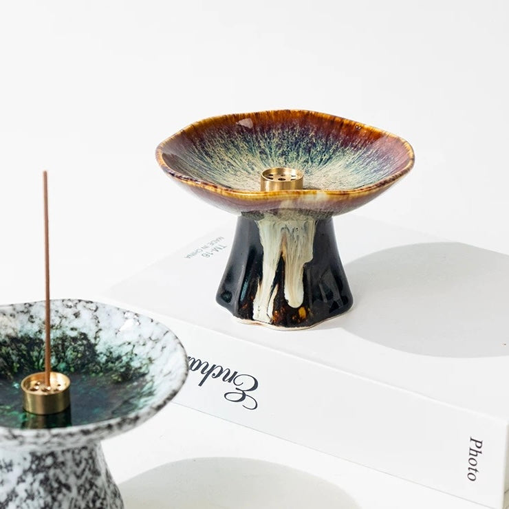 Japanese wabi-sabi style pedestal ceramic incense holders on a white background, featuring a brown-and-cream reactive glaze holder in the center and a white speckled holder with a burning incense stick in the foreground.