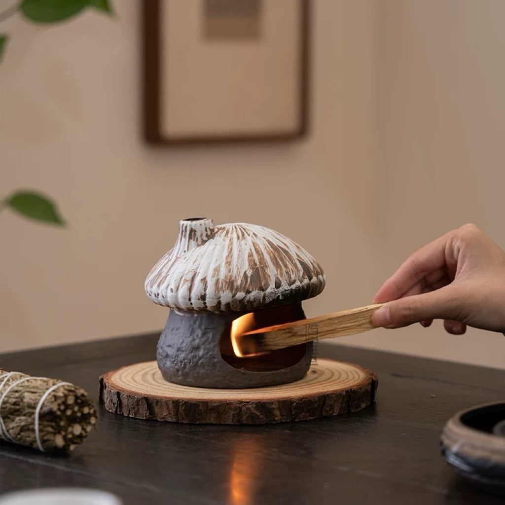 Hand placing a burning palo santo stick into a rustic ceramic mushroom house incense burner sitting on a natural wood slice coaster with sage bundle nearby