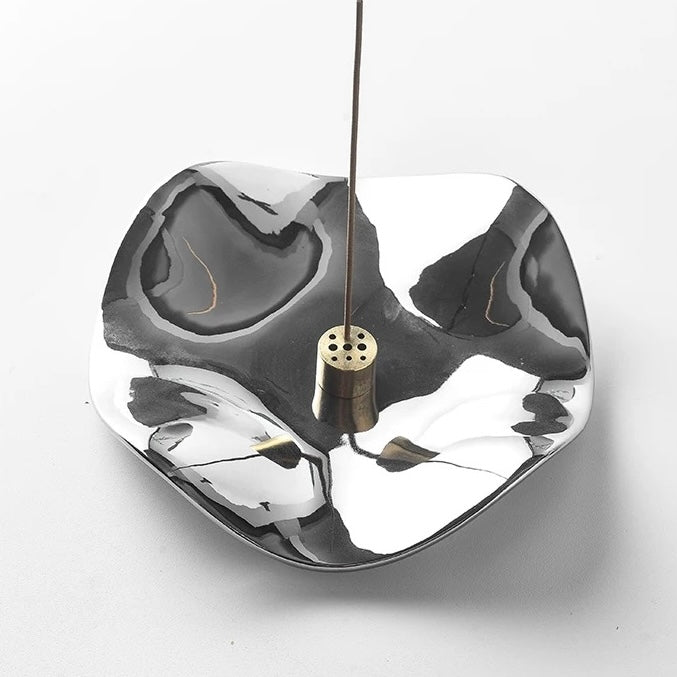 A hand placing an incense stick into the central brass holder of a silver wave-shaped tray. The image shows the interaction with the multi-hole insert.