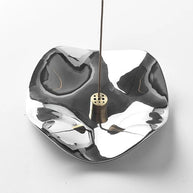 A hand placing an incense stick into the central brass holder of a silver wave-shaped tray. The image shows the interaction with the multi-hole insert.