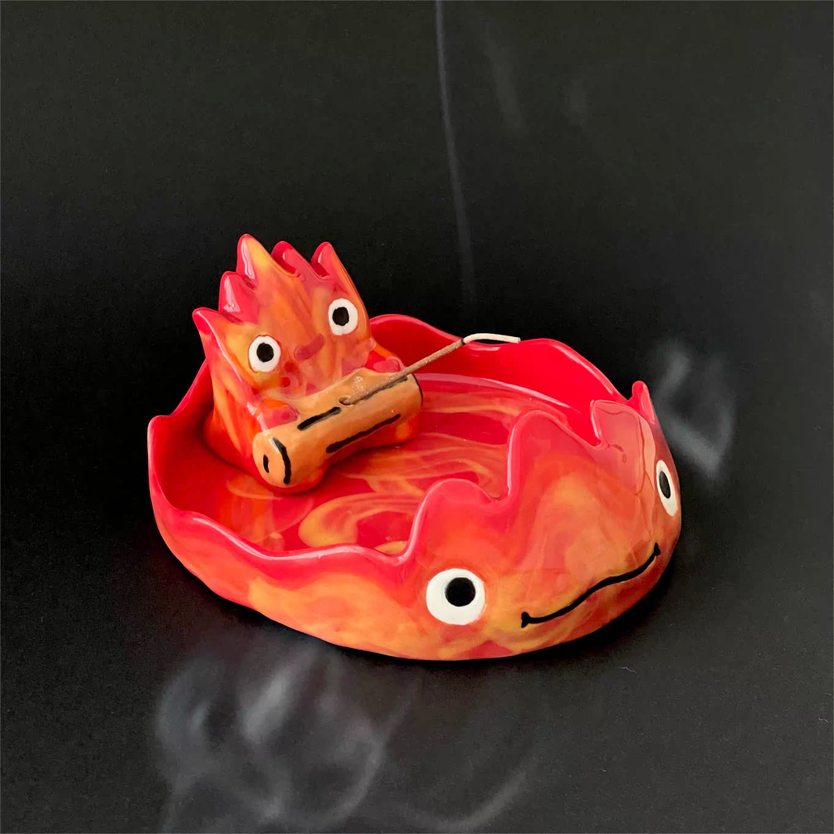 Red and orange flame character incense holder with a small log rest in a matching flame-shaped dish, faint smoke on a dark background.