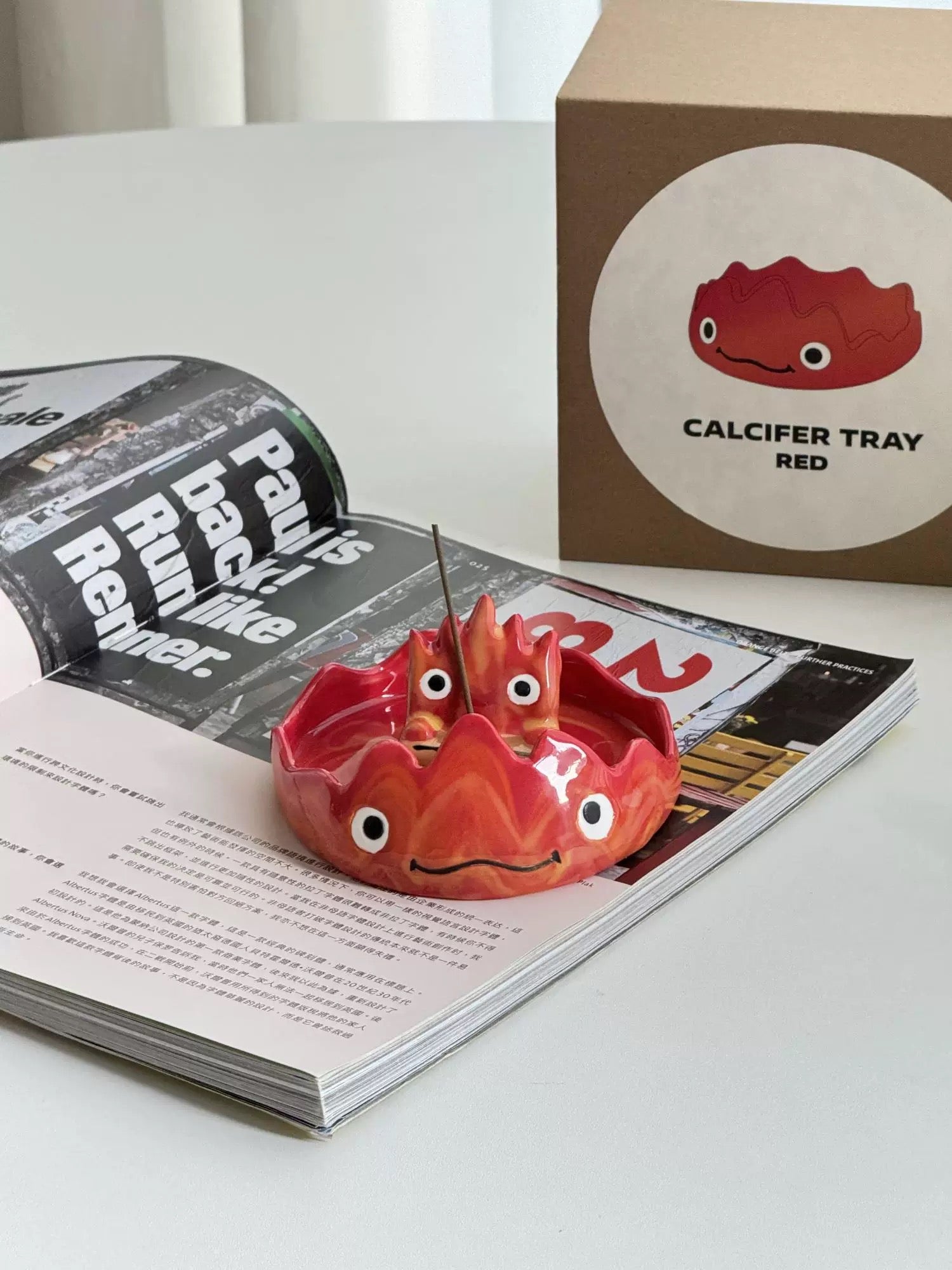 Red flame character incense holder with a matching dish and upright incense stick, placed on an open magazine.