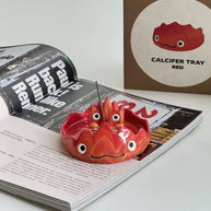 Red flame character incense holder with a matching dish and upright incense stick, placed on an open magazine.