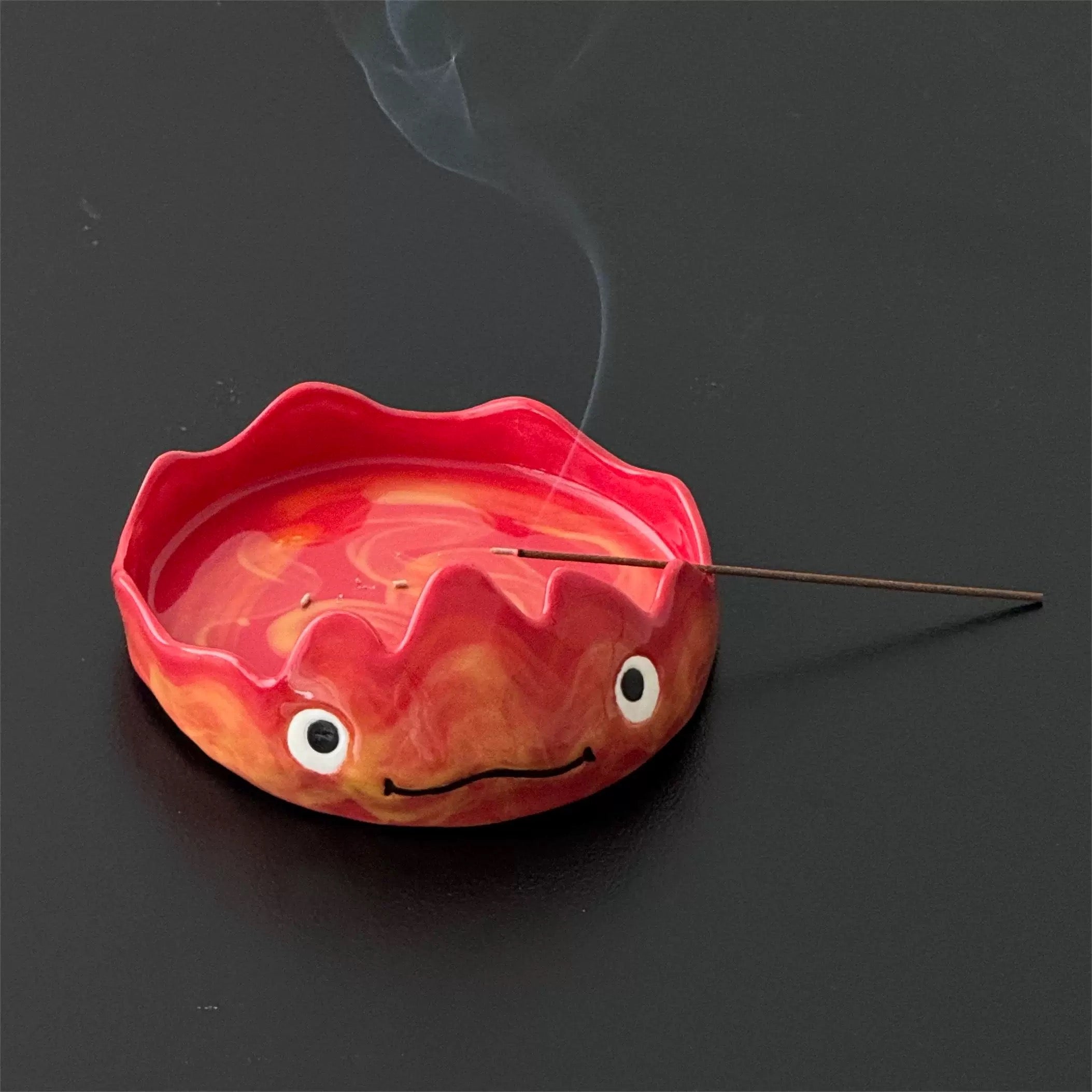 Red flame character incense tray with a lit stick resting across the rim, wisps of smoke rising on a dark background.