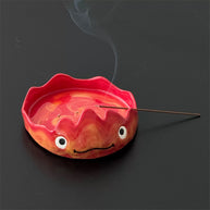 Red flame character incense tray with a lit stick resting across the rim, wisps of smoke rising on a dark background.