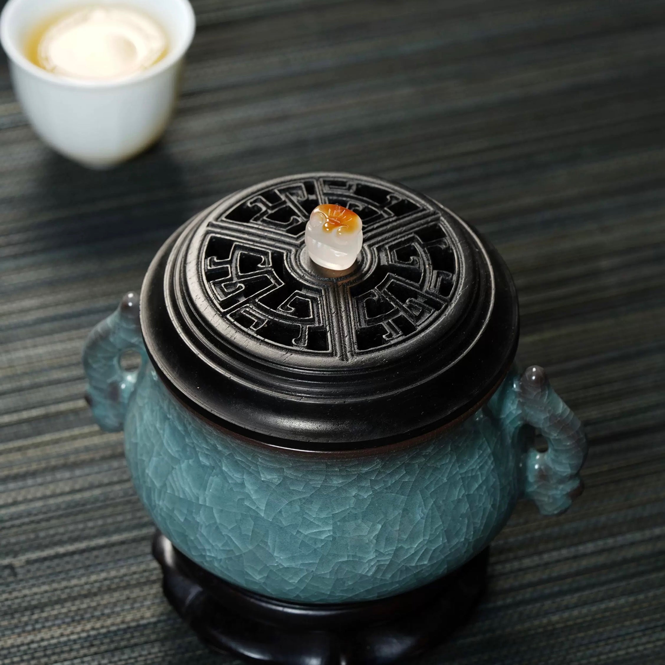 Angled view of a luxury electric incense burner with a Ge kiln ice-crackle ceramic body and carved black lid beside a teacup