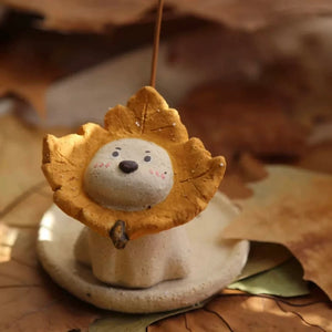 Ceramic puppy incense holder with a yellow maple leaf ruff on a round dish, incense stick inserted, autumn leaf background.