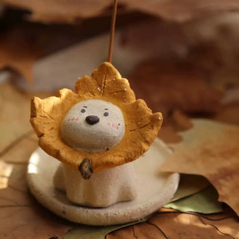 Ceramic puppy incense holder with a yellow maple leaf ruff on a round dish, incense stick inserted, autumn leaf background.