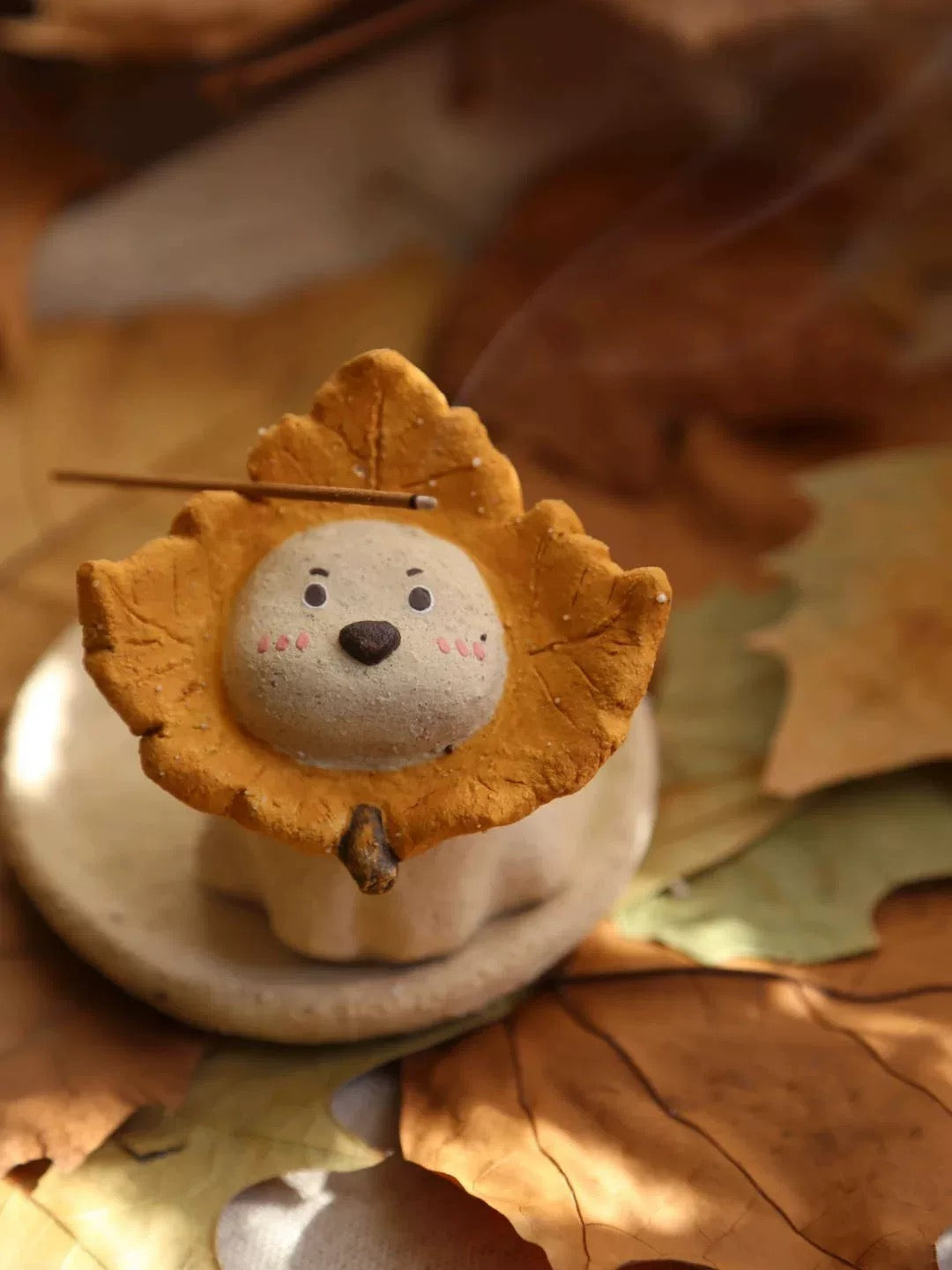 Ceramic puppy incense holder with a yellow maple leaf ruff on a round dish, incense stick resting across the top, autumn leaves background.