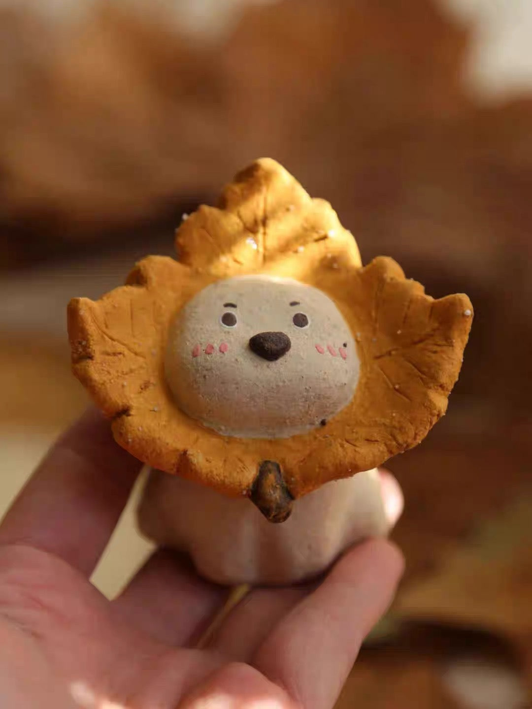 Hand holding a ceramic puppy incense holder with a yellow maple leaf ruff, close-up view against a warm autumn background.