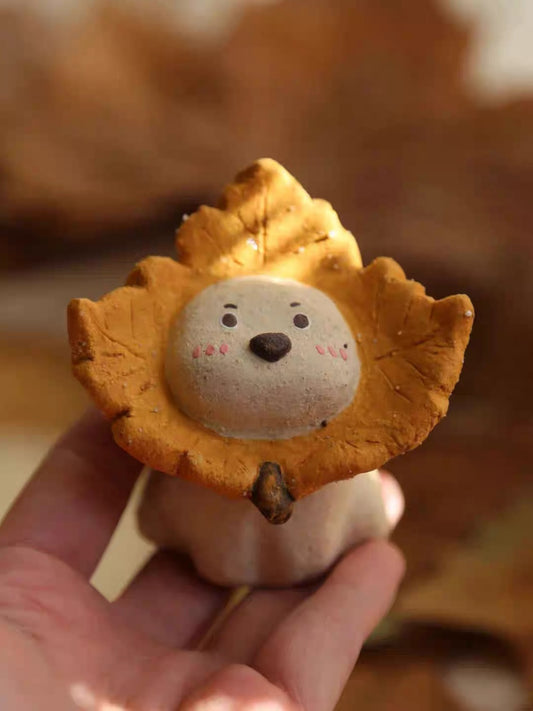 Hand holding a ceramic puppy incense holder with a yellow maple leaf ruff, close-up view against a warm autumn background.