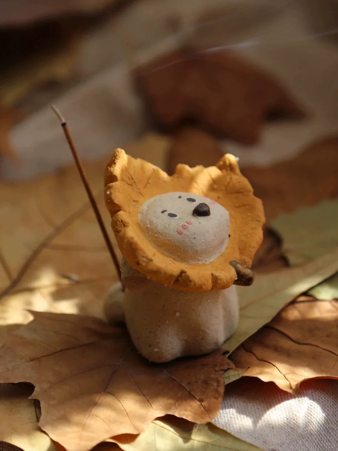Ceramic puppy incense holder with a yellow maple leaf ruff, side-inserted incense stick, set on layered autumn leaves.
