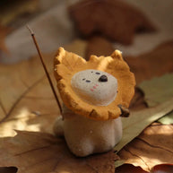 Ceramic puppy incense holder with a yellow maple leaf ruff, side-inserted incense stick, set on layered autumn leaves.