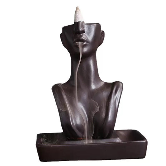 Matte black abstract face backflow incense burner with a cone incense on top, soft smoke streaming from the lips into the rectangular catch tray below on a light background.