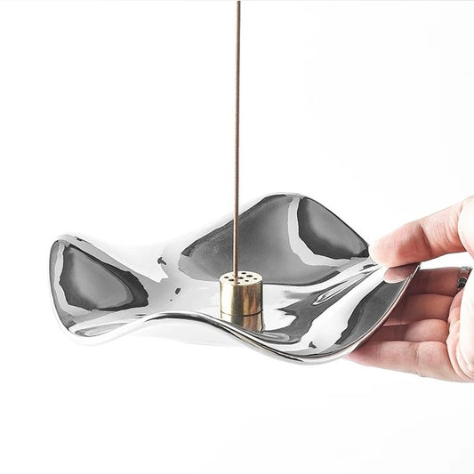 A hand holding a sculpted, fluid-shaped silver incense tray with a polished mirror finish. The tray features a central cylindrical brass holder supporting a single incense stick against a neutral background.