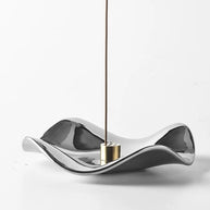 A hand is inserting an incense stick into the multi-hole brass holder sitting on a polished, fluid-shaped silver incense tray against a plain background.
