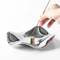 Close-up of a hand placing an incense stick into the multi-hole brass holder of a polished silver wave tray. The incense holder features a fluid, reflective design and sits on a white background.