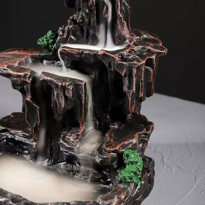 Front view of a mountain-style backflow incense burner with a lit cone at the top, white smoke cascading down dark rocky tiers into the basin below, accented with small green moss details.