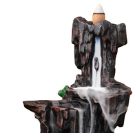 Front angled view of a mountain-style backflow incense burner with a cone placed at the top, showing white smoke flowing down the dark rocky tiers like a waterfall, with a small green moss accent on a white background.