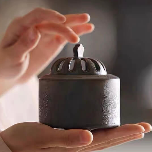 Compact obsidian black ceramic electric incense burner held between two hands to show its size and vented dome lid