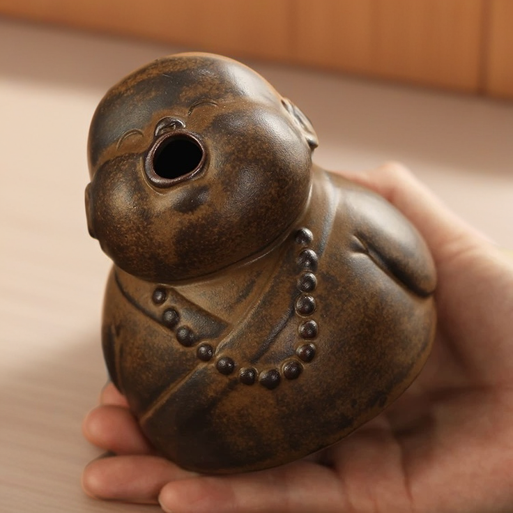 A high-angle close-up of the peaceful little monk incense burner held in a hand, highlighting the detailed brown ceramic texture, prayer beads, and the open mouth design where incense smoke emerges.