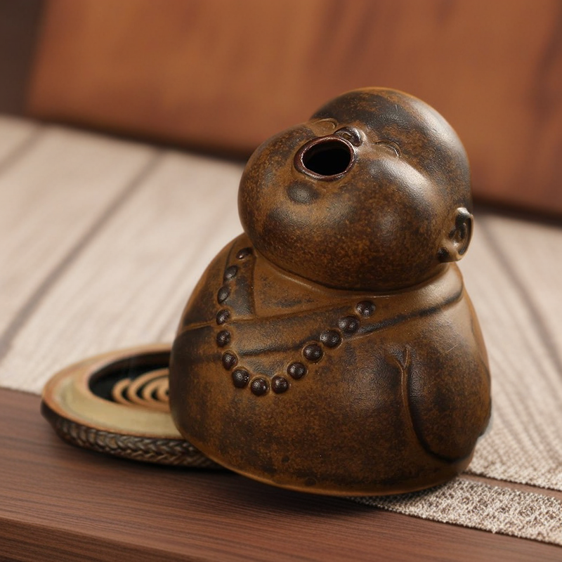 The peaceful little monk incense burner sitting on a table with the ceramic body tilted open, revealing the spiral incense coil placed on the base, showcasing the internal structure and how the incense fits inside.