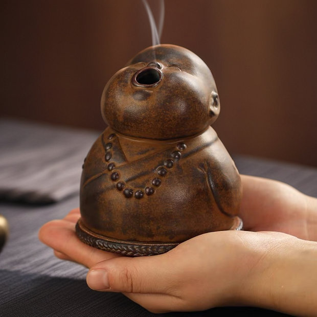 A close-up of a brown ceramic peaceful little monk incense burner held in hands, with smoke gently rising from its open mouth, featuring an authentic earthy texture and a whistling or chanting expression for zen home decor.