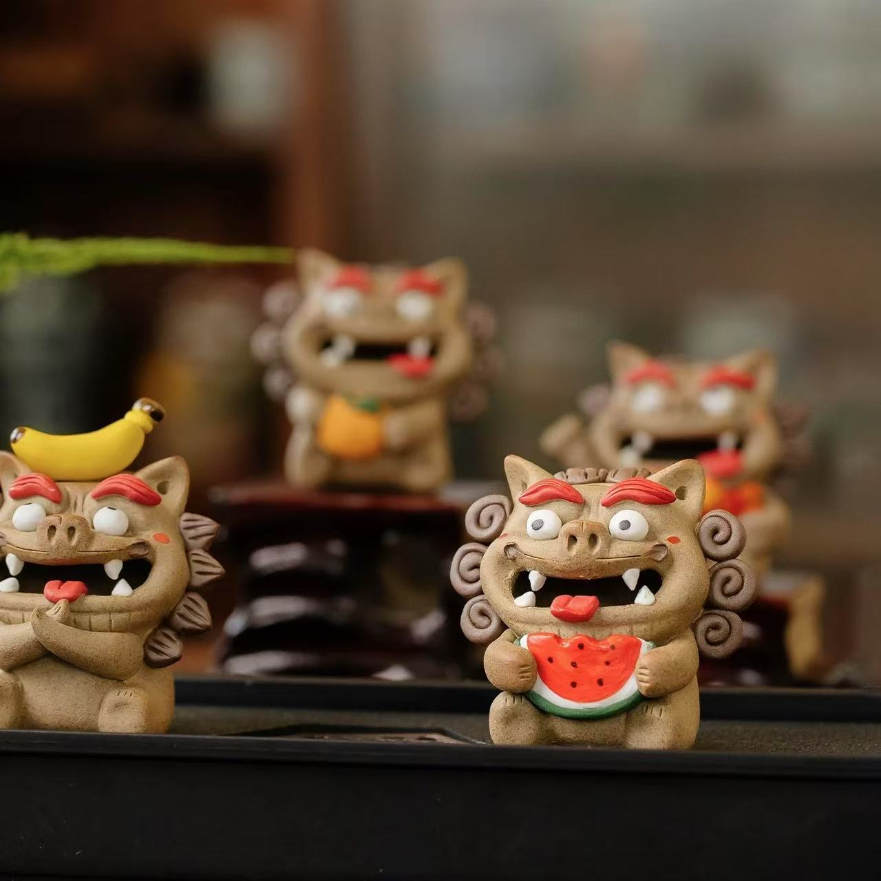 A lineup of four handcrafted ceramic Pixiu backflow incense burners on a dark tabletop, each holding a different offering (banana, watermelon, fish, orange) with wisps of smoke cascading from their open mouths.