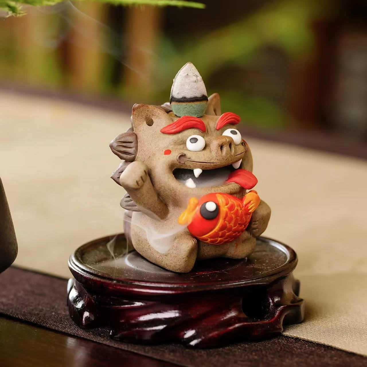 Hand‑crafted ceramic Pixiu incense burner clutching a bright red fish, with a backflow cone perched on its head and gentle smoke flowing from its open mouth, displayed on a carved wooden base.