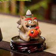 Hand‑crafted ceramic Pixiu incense burner clutching a bright red fish, with a backflow cone perched on its head and gentle smoke flowing from its open mouth, displayed on a carved wooden base.