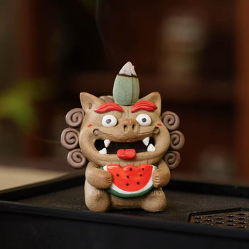 Handcrafted ceramic Pixiu incense burner holding a bright watermelon slice, with a backflow cone on its head and smoke cascading from its open mouth on a dark tray.